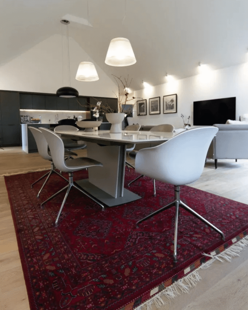 Westhill Family Home featuring an open-plan dining and kitchen area with a large white dining table and sleek modern chairs placed on a red oriental rug. The space flows into a living area with sofas and a wall-mounted TV, and features minimalist black kitchen cabinetry and pendant lighting above.