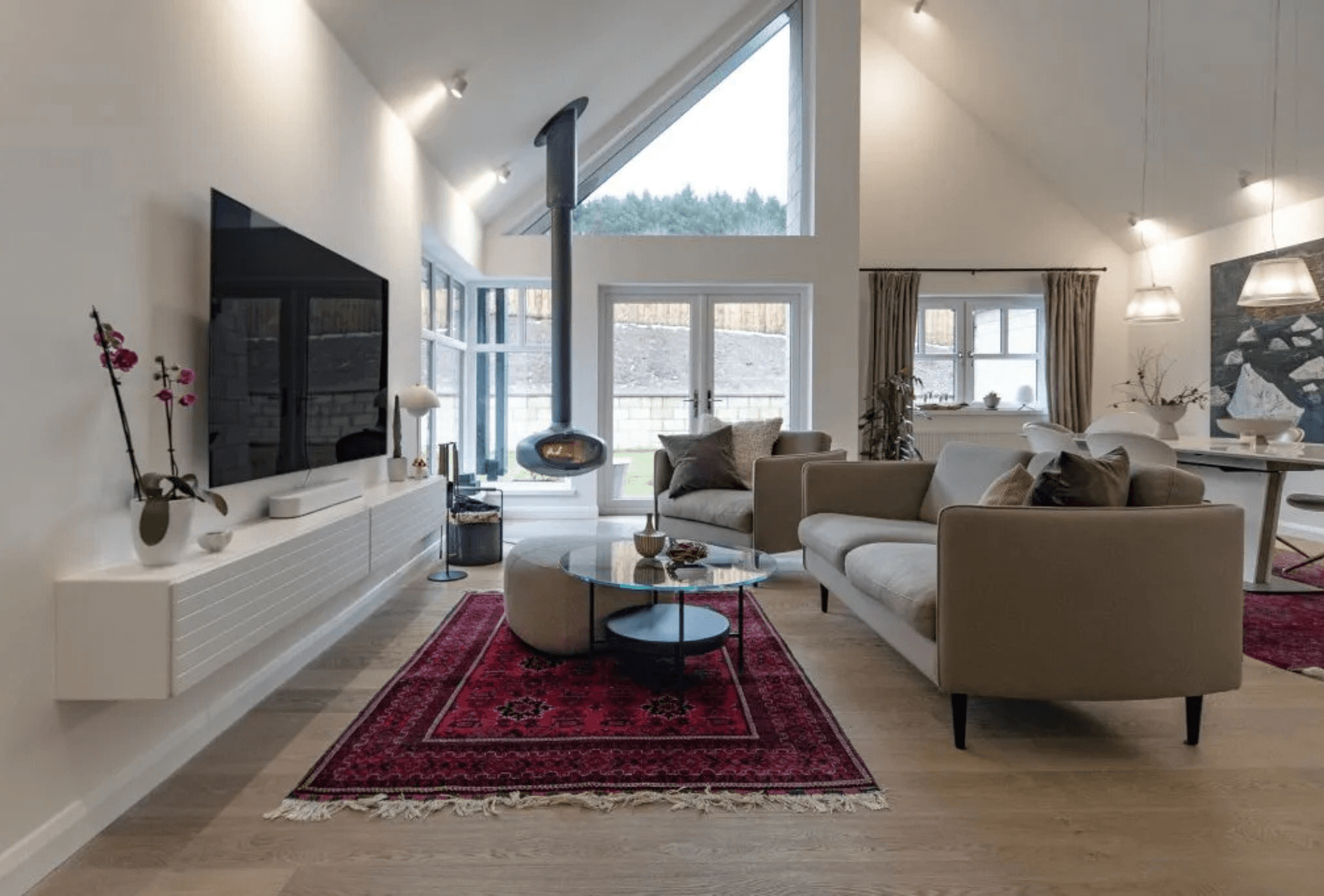 Modern living room with vaulted ceilings at the Westhill Family Home, with floor-to-ceiling windows, and a suspended black wood-burning stove, the FireBob by Firemaker. The room features a grey sofa, round glass coffee table, beige ottoman, and a bold red Persian-style rug, creating a warm yet contemporary atmosphere.