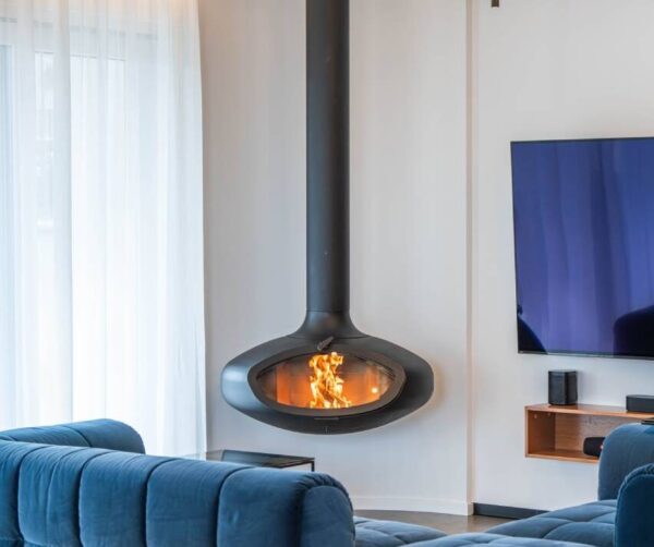 Suspended FireBob wood-burning fireplace in a contemporary living room with a blue sofa. The handcrafted circular fireplace hangs from the ceiling, creating a striking focal point while adding warmth and a calm, modern aesthetic to the space.